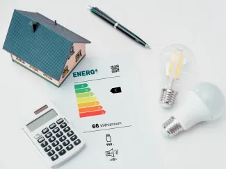 Ház, számológép, villanykörték és egy energiatakarékosságot jelző papír fehér asztalon.