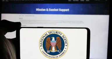 Person holding cellphone with logo of National Security Agency (NSA) on screen in front of webpage.