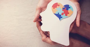 A silhouette of a person's head with a heart-shaped puzzle for a brain is held in the hands of two people.