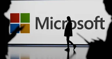 A woman is cast in silhouette walks by an illuminated Microsoft logo.