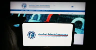 Person holding cellphone with logo of cyber security and infrastructure agency CISA on screen in front of webpage.