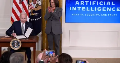 President Joe Biden sits at a table on a stage to sign an executive order while Vice President Harris applauds.