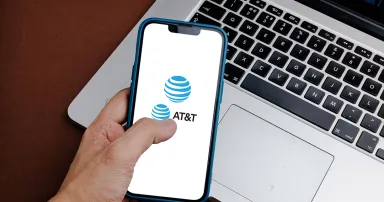 A man holds a mobile phone with the AT&T logo on the screen with a laptop in the background