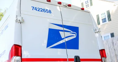 USPS sign and mailbox and truck on a sunny street corner, symbolizing reliable mail delivery and essential postal services, with a vibrant American flag in the back